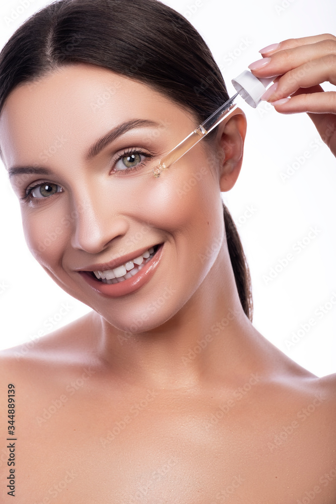 Cute smiling brunette adult girl apply make up on her fresh clean face with pipette in her hand. Beauty headshot isolated on white.