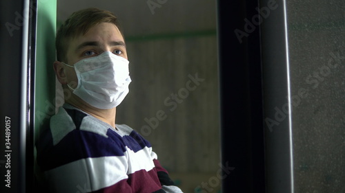 Sad young man is sitting at home in quarantine. A man sits on a window sill in a mask and looks out the window. View behind the glass.