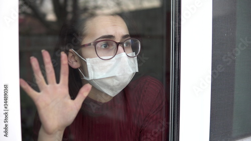 A sad young woman is sitting at home in quarantine. A girl looks out the window and puts her hand to the glass. View behind the glass.