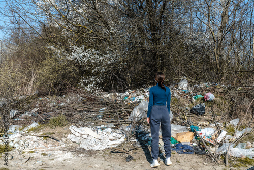 Young casual girl stands back to the camera near the trash that lying among the forest