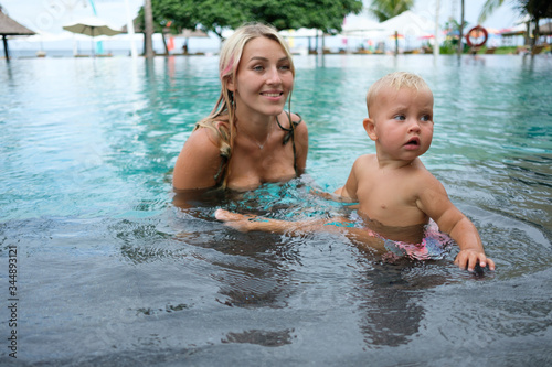Mother and baby play in outdoor swimming pool of luxury spa resort in Bali. Summer  vacation for family with children. Kids in hot tub outdoors with ocean view.