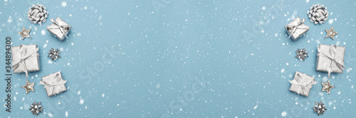 Banner of gifts or present boxes in silver with silver bows and tree ball ornaments and little silver stars, table top view, snowflakes. Composition for Christmas. Blue pastel background.