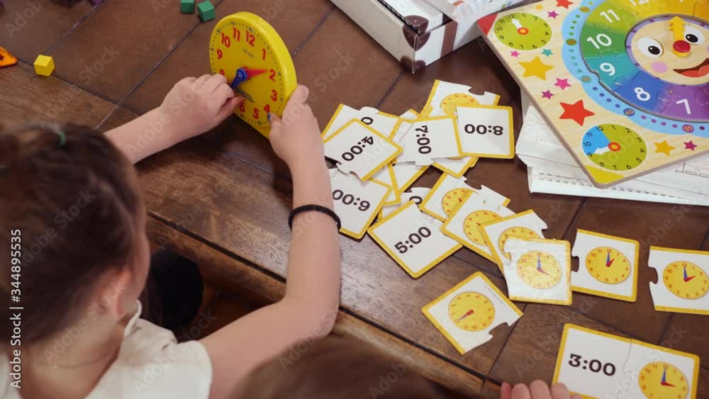 Young Kids Learning How To Tell Time Using A Toy Clock And Illustrations. High angle video showing children learning how to tell time at home during pandemia time.
