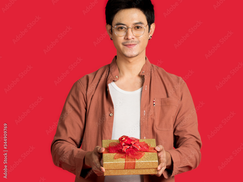 Portrait of handsome Asian young man holding golden gift box with red ribbon, wearing casual clothing and eyeglasses isolated on red background. Portrait of handsome Asian young man holding golden gift box with red ribbon, wearing casual clothing and eyeglasses isolated on red background.