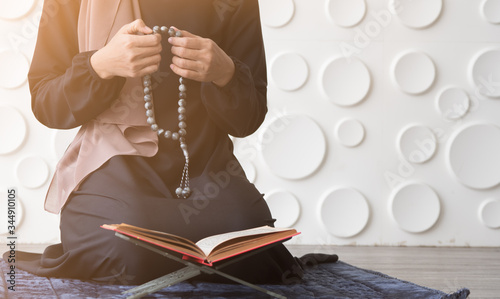 Close up hand holding rosary beads, praying or reading quran or kuran on the small table while sitting on rug or carpet on white pattern background.