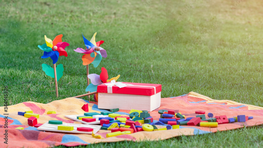 Fototapeta premium Colorful wooden blocks on the rug next to opened gift box and rainbow pinwheels on the grass in the garden or park.
