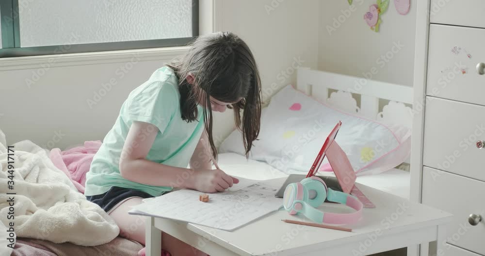 young girl home schooling or doing homework in her bedroom. She is writing in a notepad at a small table with a tablet computer and headphones on the table. She flicks her hair