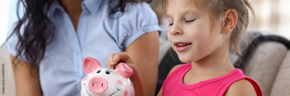 Smiling little girl making wish while putting coin in piggybank slot Smiling little girl making wish while putting coin in piggybank slot