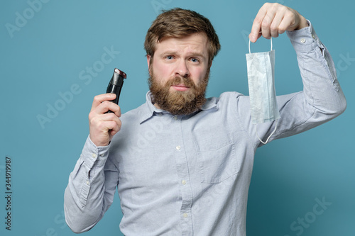 Man with a trimer in hands is upset because he has to shave beard to wear a medical mask. Concept of quarantine during a pandemic. photo