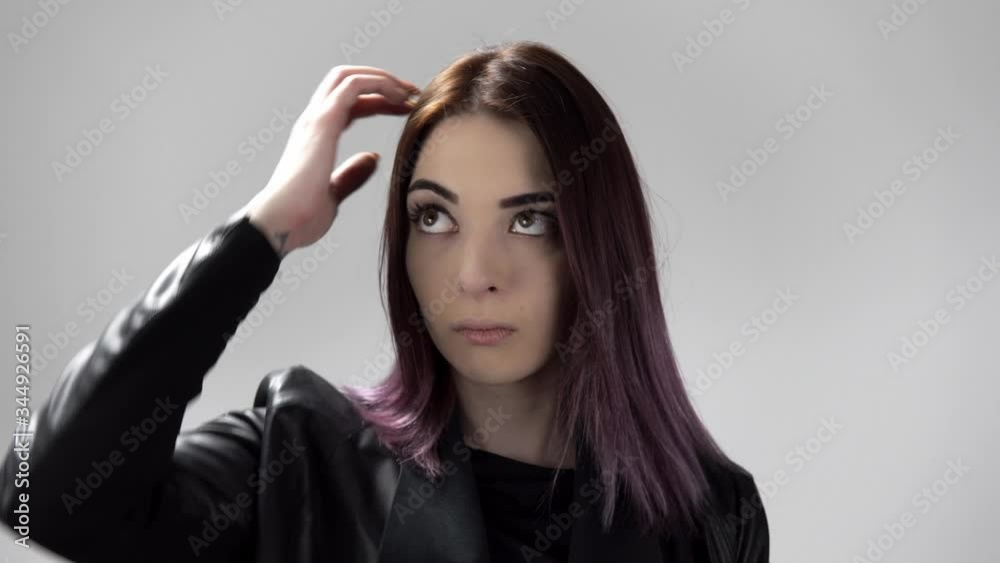 Portrait of young woman with brown eyes thinking hard and scratching her head. The girl dressed in black leather jacket is standing in the studio with white background. Concept of showing emotions. Portrait of young woman with brown eyes thinking hard and scratching her head. The girl dressed in black leather jacket is standing in the studio with white background. Concept of showing emotions.