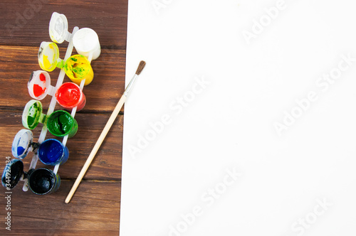 a set of colorful paints in jars and a paint brush on a wooden table on a white sheet of paper top view