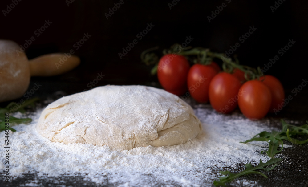homemade pizza dough or pies on a dark rustic background, next to a branch of tomatoes and arugula greens and a rolling pin homemade pizza dough or pies on a dark rustic background, next to a branch of tomatoes and arugula greens and a rolling pin