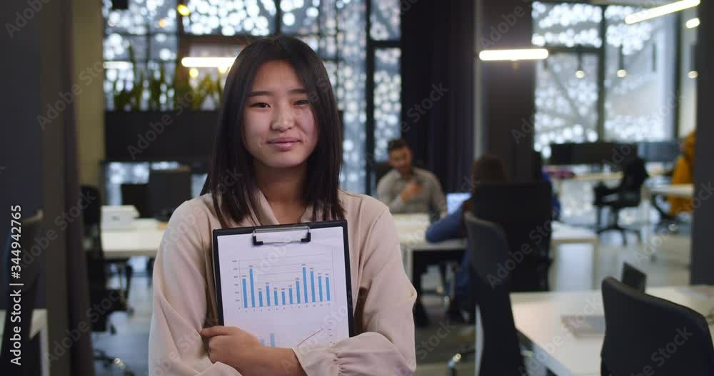 Portrait of young Asian beautiful woman standing in company office and looking at camera. Pretty girl holding some papers with charts and graphics. Female worker of firm. Portrait of young Asian beautiful woman standing in company office and looking at camera. Pretty girl holding some papers with charts and graphics. Female worker of firm.