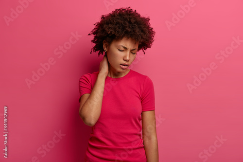 Displeased dark skinned woman touches neck, feels pain in back after falling from stairs, suffers from spine painful feelings, tilts head and closes eyes, wears casual t shirt, isolated on pink wall