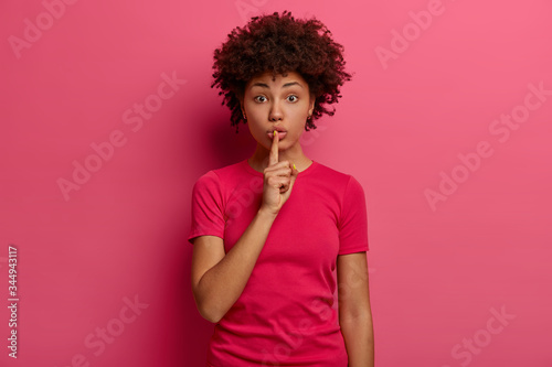Photo of shocked surprised dark skinned woman keeps index finger over lips  makes silence gesture  tells big secret  looks mysteriously at camera  asks keep quiet  dressed casually. Hush  dont speak