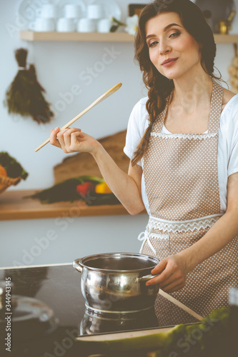 Young brunette woman cooking soup in kitchen. Housewife holding wooden spoon in her hand. Food and health concept