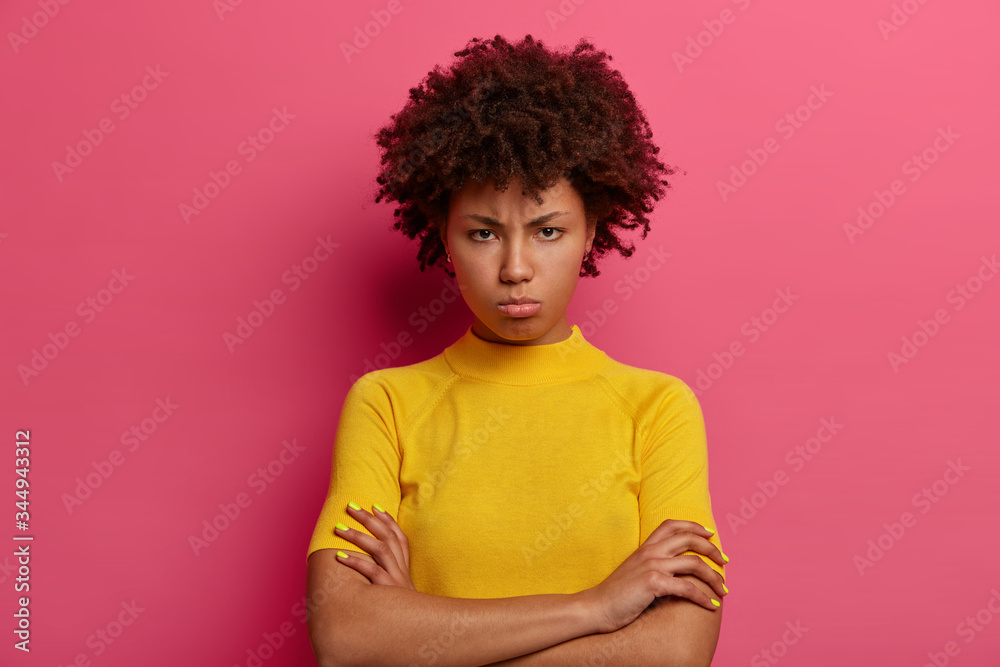Discontent woman with sulking expression, keeps arms folded, looks angrily at camera, offended by hearing bad words, frowns face in displeasure, wears bright yellow clothes, isolated on pink wall Discontent woman with sulking expression, keeps arms folded, looks angrily at camera, offended by hearing bad words, frowns face in displeasure, wears bright yellow clothes, isolated on pink wall