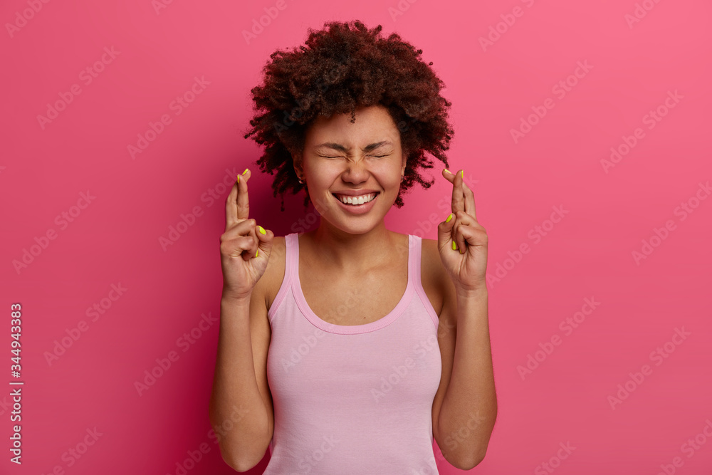 Joyful pleased cute woman stands with crossed fingers, wants dream come true, believes in good luck, wears casual vest, makes wish, awaits positive reply or good news, stands hopeful indoor. Joyful pleased cute woman stands with crossed fingers, wants dream come true, believes in good luck, wears casual vest, makes wish, awaits positive reply or good news, stands hopeful indoor.