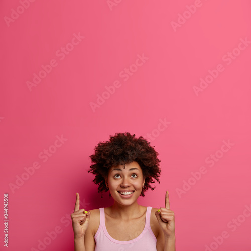 Positive ethnic woman points fingers up at copy space above head for your advertisement, attracts attention to great offer, smiles happily, isolated on pink background, gives advice, promots product
