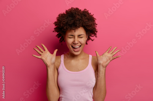 Positive dark skinned woman laughs and raises palms, feels very glad, canot stop laughter, closes eyes and giggles, wears casual vest, isolated on pink background. Positive emotions and feelings