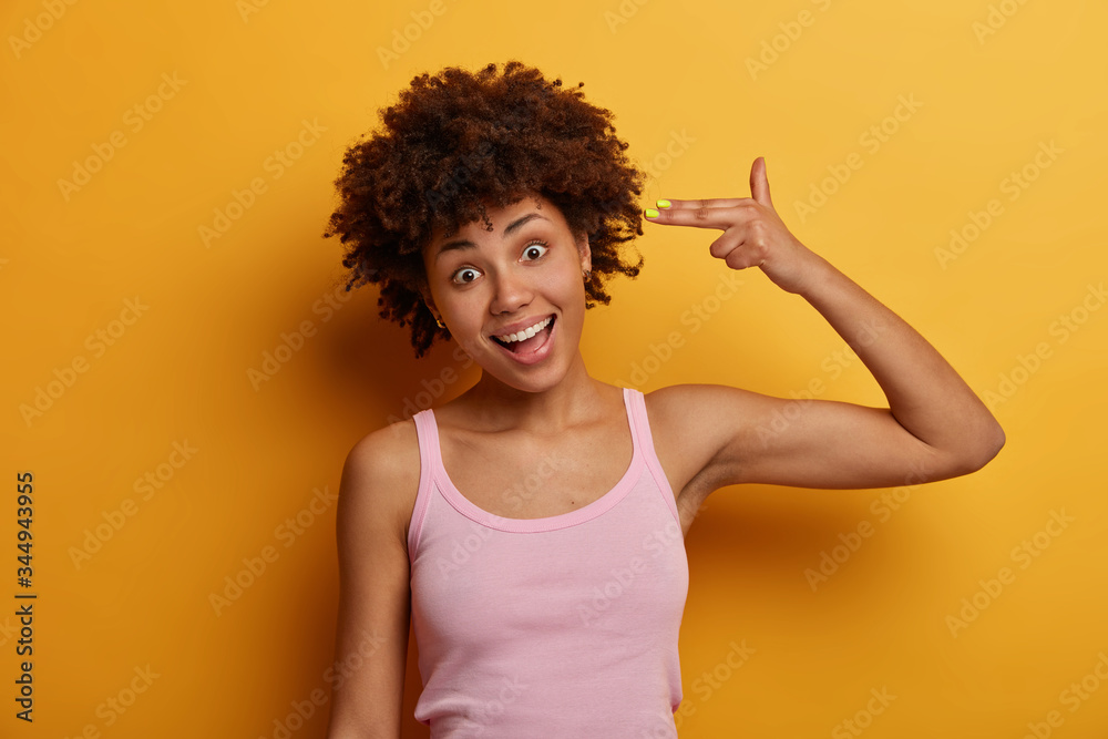 Portrait of positive teenage girl makes suicide gesture, shoots in temple with fingers, gazes happily at camera, wears casual t shirt, feels bored, tries to amuse herself, stays at home, isolated Portrait of positive teenage girl makes suicide gesture, shoots in temple with fingers, gazes happily at camera, wears casual t shirt, feels bored, tries to amuse herself, stays at home, isolated