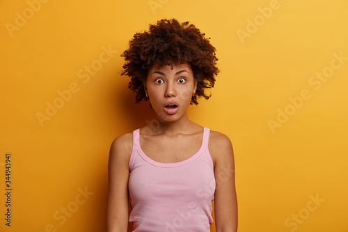 Astonished African American woman realizes lie from close person, looks at camera with shocked face expression, opened mouth, stands in stupor against yellow background. Human reaction and emotions