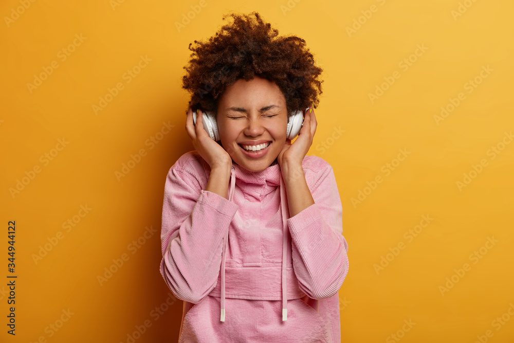 Joyful curly haired woman enjoys every bit of favorite song, listens music in stereo headphones, closes eyes and smiles broadly, inspired with awesome music, dressed in casual wear, stands indoor Joyful curly haired woman enjoys every bit of favorite song, listens music in stereo headphones, closes eyes and smiles broadly, inspired with awesome music, dressed in casual wear, stands indoor