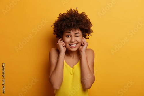 Indoor shot of beautiful woman plugs ears, avoids very loud music on party, winks eye, clenches teeth, dressed in yellow clothes, poses indoor, cannot listen noise, avoids very loud neighbours