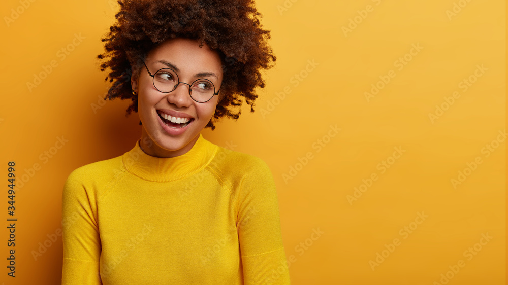 Studio shot of good looking woman laughs as looks right, reads funny promo on blank space, has satisfied smile, wears vivid yellow jumper, feels happy and secure. People, emotions, beauty concept Studio shot of good looking woman laughs as looks right, reads funny promo on blank space, has satisfied smile, wears vivid yellow jumper, feels happy and secure. People, emotions, beauty concept