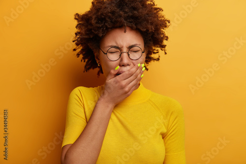 Depressed dark skinned woman cries from despair, covers mouth, looks miserable, whins loudly, wears optical glasses, yellow casual t shirt, poses indoor, tired of problems. Negative emotions © wayhome.studio 