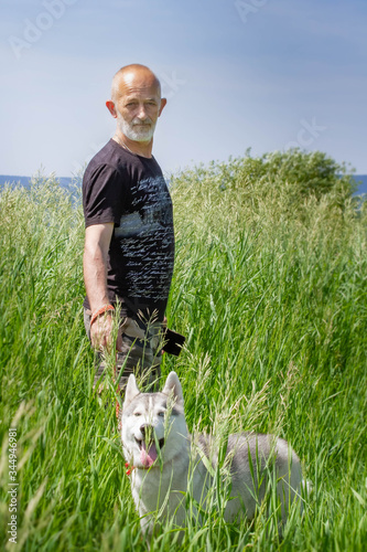 An old man and sled dog walk near the river. Alpine landscape. Active leisure pensioner. An elderly man is smiling. Walk with Siberian Husky.