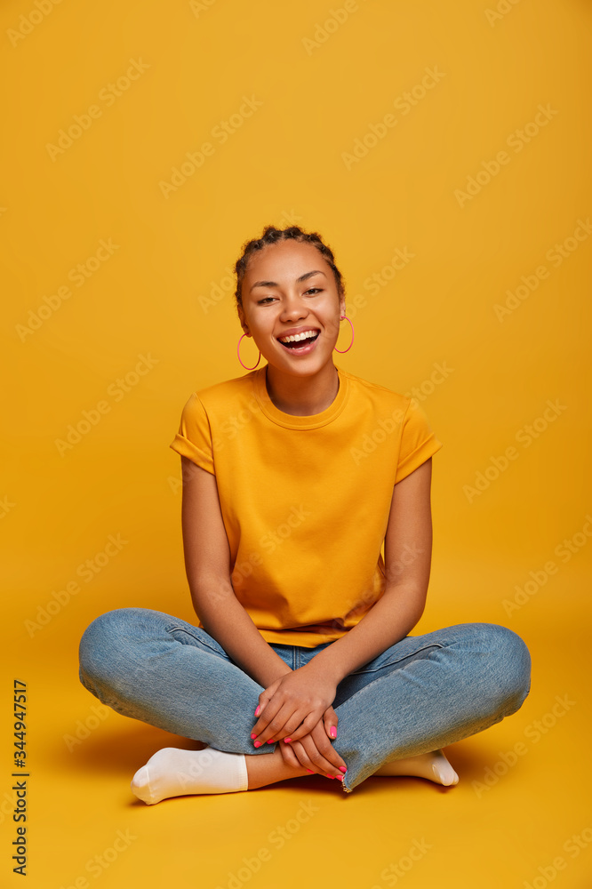 Relaxed beautiful millennial woman sits crossed legs, laughs, has hilarious conversation, wears t shirt, jeans, socks, has cornrow hairstyle, isolated on yellow background, expresses carefree emotions Relaxed beautiful millennial woman sits crossed legs, laughs, has hilarious conversation, wears t shirt, jeans, socks, has cornrow hairstyle, isolated on yellow background, expresses carefree emotions