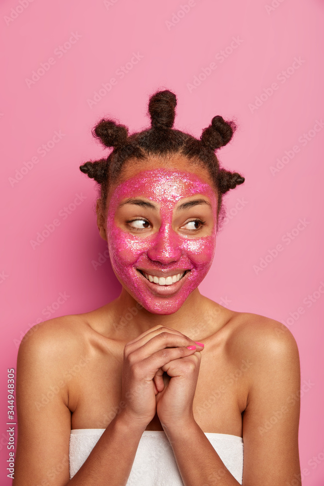 Studio shot of cheerful beauty model stands wrapped in towel, applies nourishing mask, keeps hands pressed together, looks aside, poses over rosy studio wall. Face care, wellness, cosmetology concept