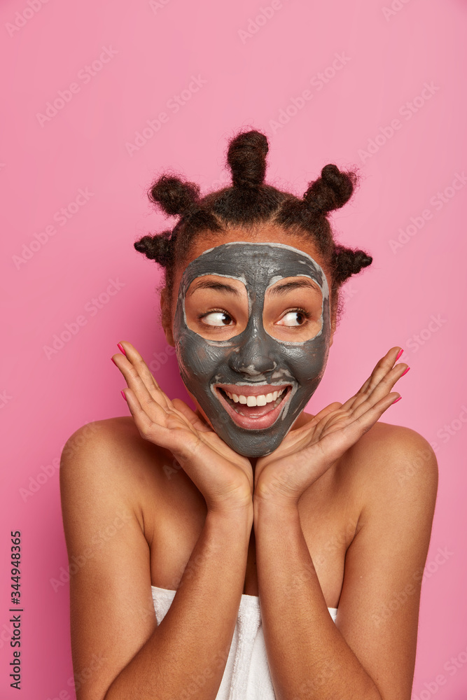 Skin care, cosmetology and well being concept. Positive dark skinned model spreads palms over face, applies moisturizing mask for cleaning skin, has beauty procedures after bath, wears towel over body
