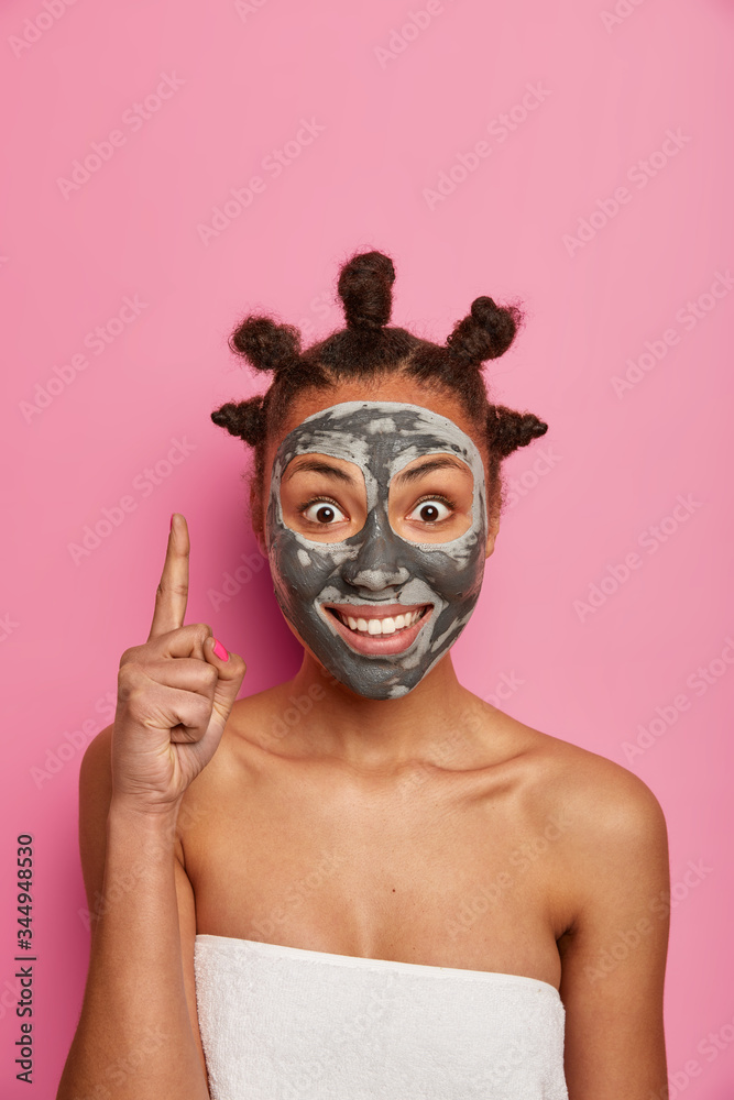 Positive Afro American woman points above with index finger, cares about skin, applies clay mask, advertises nice beauty product, cares about appearance, looks young and beautiful, poses indoor