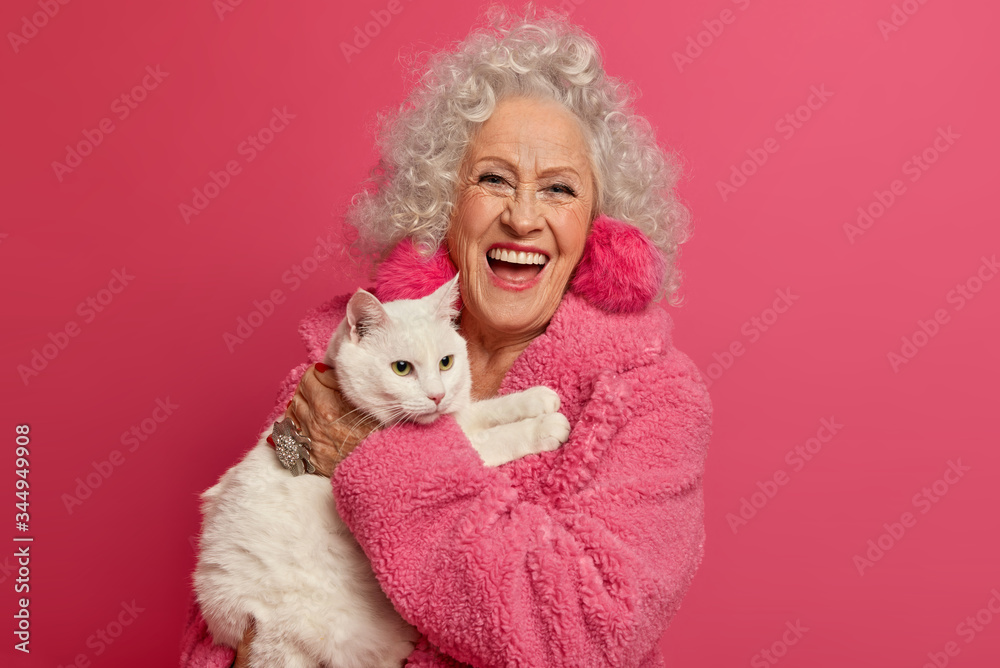 Old woman pet lover ready for walk with favorite cat, laughs happily, expresses positive emotions, going to vet clinic, cares about cat, wears rosy warm gown, stands indoor. Mature people, animals