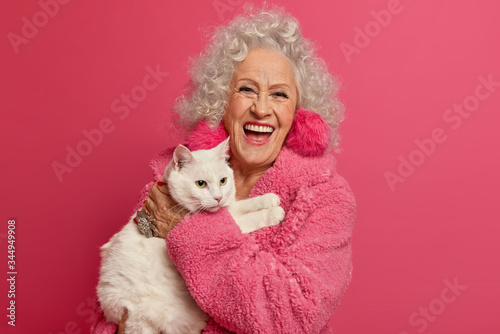 Old woman pet lover ready for walk with favorite cat, laughs happily, expresses positive emotions, going to vet clinic, cares about cat, wears rosy warm gown, stands indoor. Mature people, animals