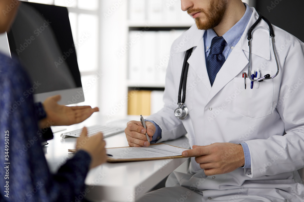 Unknown red-bearded doctor and patient woman discussing current health examination while sitting in sunny clinic, close-up. Medicine concept