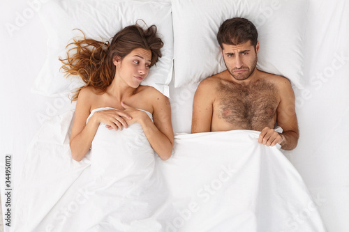 Man impotent has erectile dysfunction, looks under blanket with frustrated expression, unsatisfied woman lies near, have relationship problem because of sex failure. Mens health and impotence concept photo