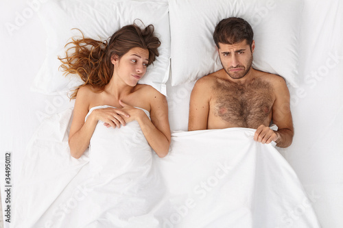 Man impotent has erectile dysfunction, looks under blanket with frustrated expression, unsatisfied woman lies near, have relationship problem because of sex failure. Mens health and impotence concept