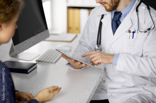 Unknown red-bearded doctor and patient woman discussing current health examination while sitting and using tablet computer in clinic, close-up. Medicine concept