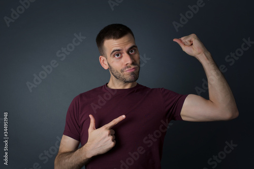 Waist up shot of blond man raises hand to show his muscles, feels confident in victory, looks strong and independent, smiles positively at camera, stands against gray background. Sport concept.