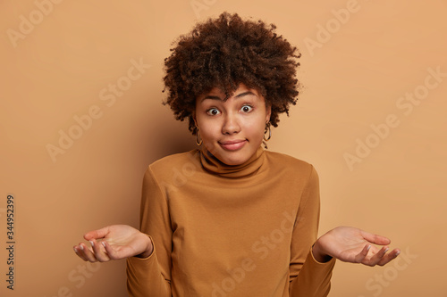 Confused dark skinned adult female has question, feels indecisive and hesitant, shrugs shoulders, poses unaware, being confused, cant help, wears brown turtleneck, isolated on beige background