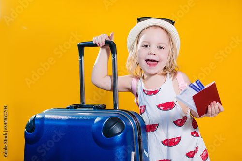 child girl on a yellow background with a blue suitcase in a hat and a summer dress with tickets, going on a trip, summer holidays, borders opened, travel in children