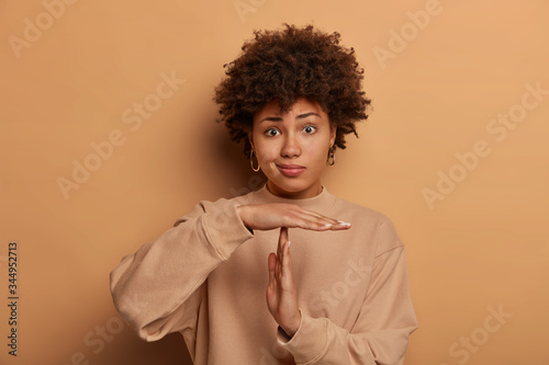 Confused dark skinned woman makes pause or break time gesture, timeout sign, wears sweatshirt, stands against brown background, asks to wait, upset about deadline. Body language concept. Time limit photo