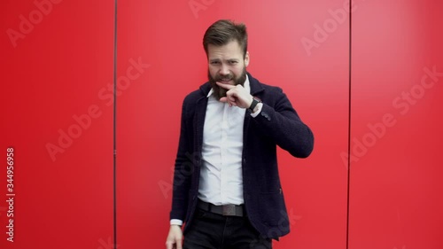Brunette bearded man in casual clothes is actively gesturing and explaining something on the camera while standing on the red background. Hyperactive young guy is dressed in white shirt and dark