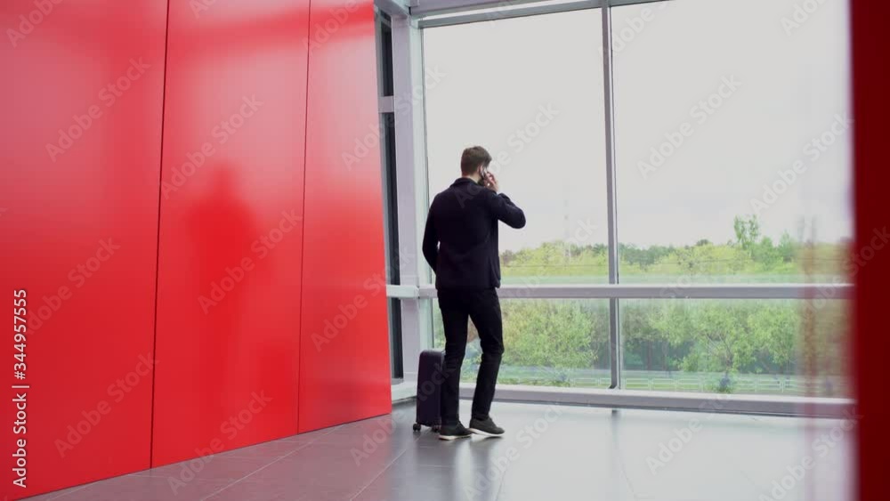 Bearded businessman enters the room with red walls while talking on the phone and then he is standing with suitcase on the wheels near panoramic windows. Picture windows overlook park with green trees