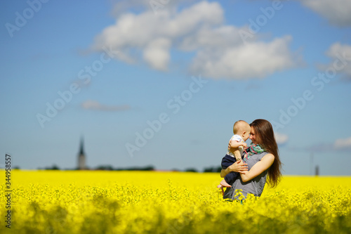 Young mother holding baby in hands in bright yellow flower field in sunny summer day, bright blue sky and yellow meadow © Vladimir