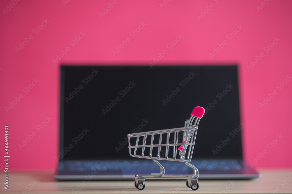 Online shopping concept. Mini grocery cart and laptop on a pink background. Small cart and personal computer.