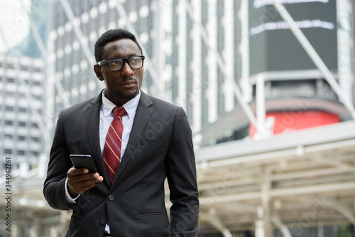 Young handsome African businessman holding mobile phone in front of modern building at Bangkok city